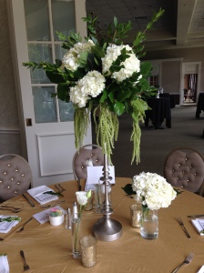 The tall centerpiece with hydrangeas and a lush bedding of greens surrounded by  vases with flowers or candles  